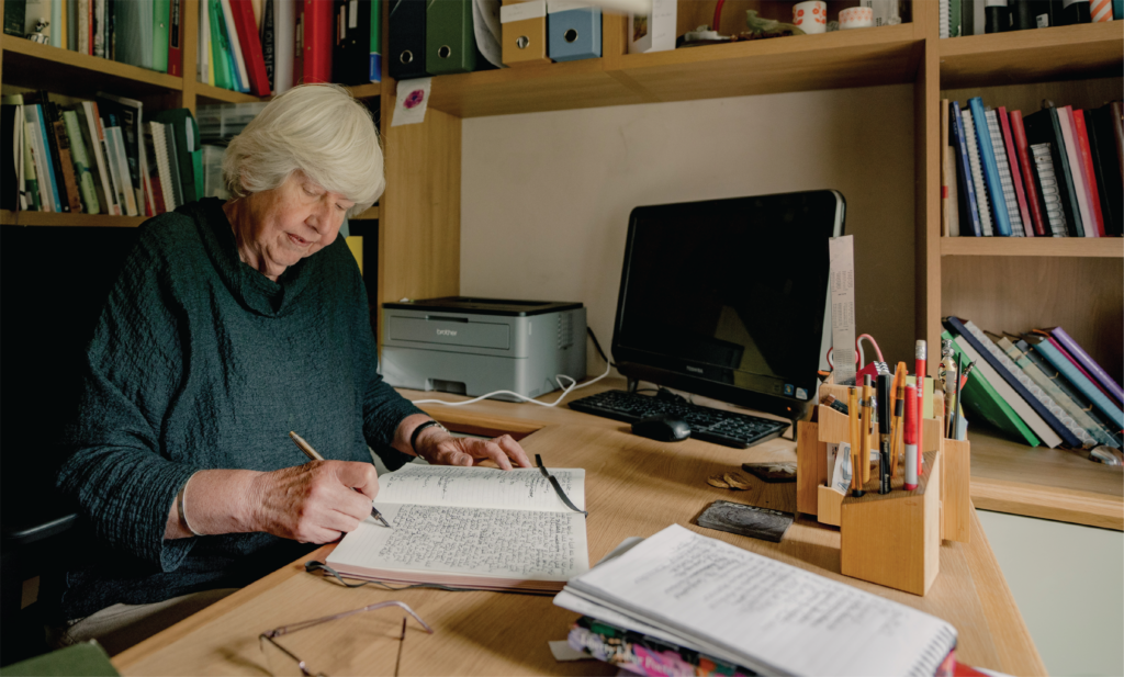 Anne Youngson writes in a notebook beside her computer at a wooden desk, surrounded by shelves of books.